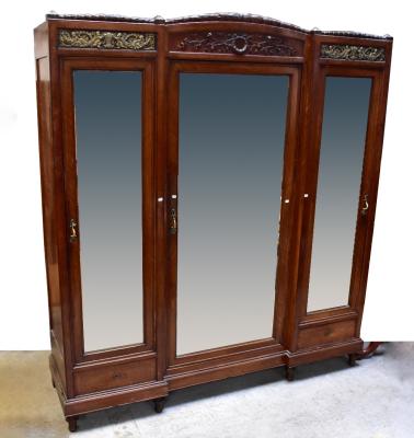 An early 20th century French Empire Revival walnut and kingwood two-piece bedroom suite, comprising a large triple wardrobe with carved pediment above a large bevelled mirrored door, flanked by a pair of bevelled mirrored doors, surmounted with gilt brass cherubic panels, above a single base drawer, raised on turned supports, 238 x 213 x 60cm, and a matching marble-topped dressing chest with bevelled mirror plate above inset marble panel, above three frieze drawers and three long drawers, raised