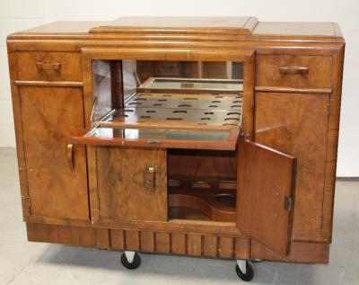 A c1950s Art Deco style mahogany sideboard/cocktail cabinet, stepped top above clear glass fall-front cocktail unit with mirror back, two frieze drawers either side above two cupboard doors, each side with small frieze drawer and cupboard door to shaped plinth base, 135 x 103cm.