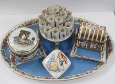 An early 20th century Dresden oval hand-painted table centre for cigarettes, with central cylindrical holders for approximately twenty cigarettes, two hand-painted ashtrays, a French-style perfume bottle painted with figures in period costume, a Limoges-style trinket pot in the form of a treasure chest and a commemorative hand-painted trinket pot for Paris, L'Arc de Triomphe (4).